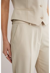 Close-up van een persoon die beige op maat gemaakte broek en bijpassend vest draagt, met de linkerhand langs het lichaam en een zilveren ring aan.