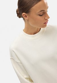 Sudadera blanca con cuello redondo, corte relajado y puños acanalados. Textura suave, con detalles de costura visibles a lo largo de las costuras.