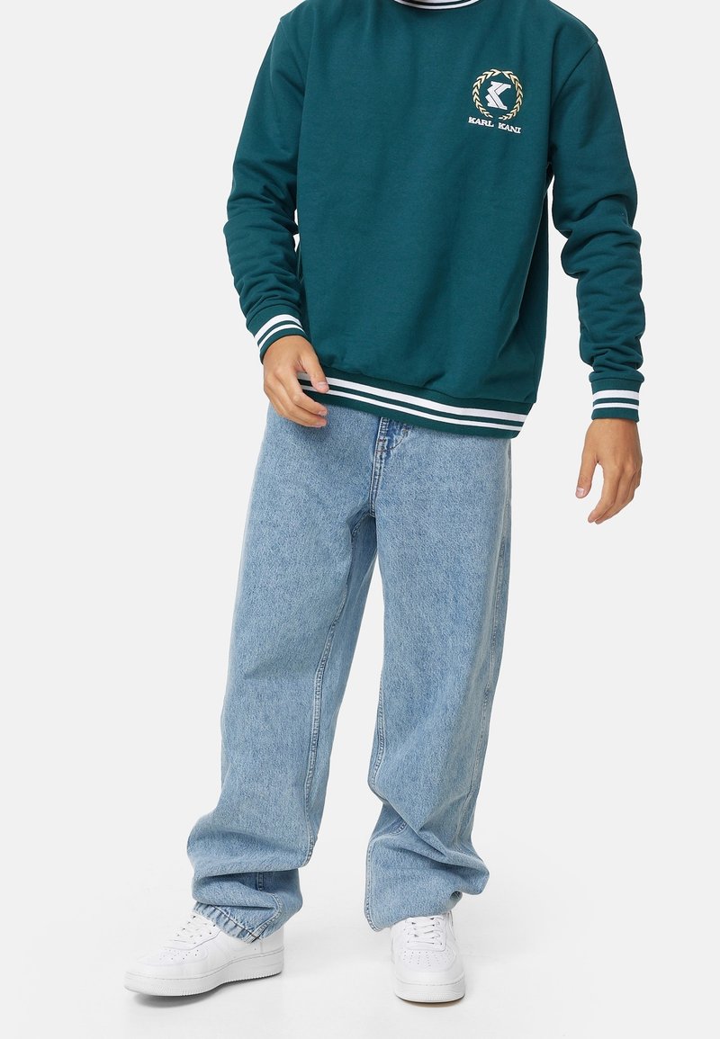 Felpa crewneck color teal con strisce bianche, con logo sul petto. Abbinata a jeans baggy blu chiaro e sneakers bianche.