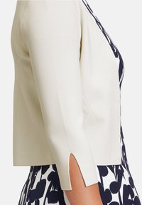 Knit cream cardigan with three-quarter sleeves, featuring side slits. Worn over a navy patterned dress with bold white shapes.