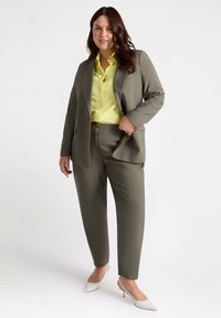 Olive green tailored suit with a single-breasted jacket, paired with a bright yellow button-up shirt. White pointed-toe heels complete the look.