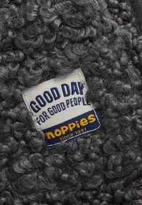 Szary materiał teksturowany z puszystą, kręconą powierzchnią, z naszywką, na której widnieje napis: "DOBRY DZIEŃ DLA DOBRYCH LUDZI" oraz "noppies od 1991 roku."