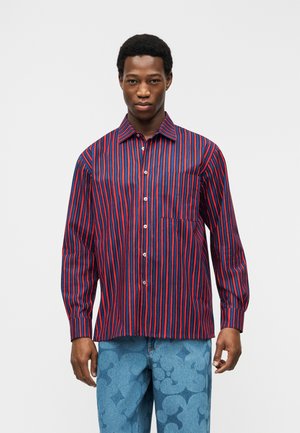 Muž nosiaci košeľu s dlhým rukávom zapínanú na gombíky s červenými a námorníckymi zvislými pruhmi a svetlomodré rifle s kvetinovým vzorom, stojaci pred bielym pozadím.