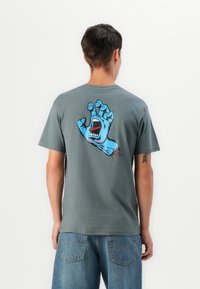 Person med grå t-shirt med blå skrigende h�åndgrafik og "Santa Cruz" tekst på ryggen, sammen med løse denimshorts.