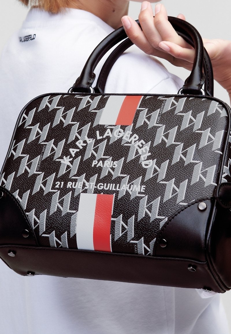 Main tenant un sac à main noir Karl Lagerfeld avec motif blanc « KL », rayures verticales rouges et blanches, et texte d'adresse Paris.
