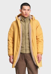 Jeune homme portant un manteau d'hiver jaune à capuche avec doublure polaire par-dessus une veste polaire verte zippée et un pantalon marron sur un fond uni.