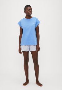 Blauwe katoenen T-shirt met korte mouwen en geborduurde tekst, gecombineerd met gestreepte shorts. Model staat op blote voeten tegen een effen achtergrond.