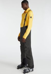 Pantalones de esquí negros con tirantes ajustables, combinados con una camiseta de manga larga amarilla. Los pantalones cuentan con bolsillos y una textura suave y resistente al agua.