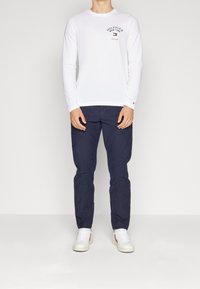 Weißes Langarm-T-Shirt mit Rundhalsausschnitt und gesticktem Logo, kombiniert mit marineblauen Hosen und weißen Sneakers mit roten Akzenten.