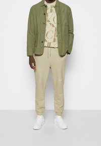 Olivgrön jacka, beige grafisk t-shirt med bokstäver, beige sweatpants med uppvikta anklar och vita sneakers med minimal detaljering.