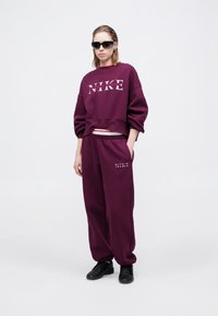 Persona che indossa un felpa Nike viola e pantaloni della tuta coordinati, scarpe nere e occhiali da sole scuri, in piedi contro uno sfondo bianco.