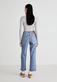 Jean bleu clair taille haute avec une jambe droite, doté d'un patch en cuir à l'arrière, associé à un haut gris à manches longues et des baskets marron.