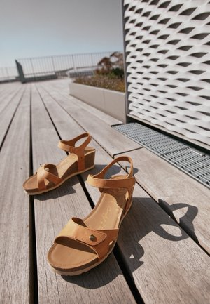 Las sandalias de cuña de cuero marrón presentan correas cruzadas, una correa alrededor del tobillo, una plantilla texturizada y una suela tipo corcho con un detalle decorativo de hardware.