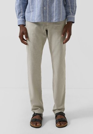 Mann trägt hellbeige Hose, ein blau gestreiftes Langarmhemd und schwarze offene Zehensandalen, steht vor einem einfarbigen Hintergrund.
