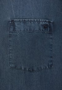 Niebieska tkanina dżinsowa posiada prostokątne tylne kieszenie z przeszywanym brzegiem oraz minimalnym strzępieniem w górnym rogu. Gładka tekstura z delikatnym połyskiem.