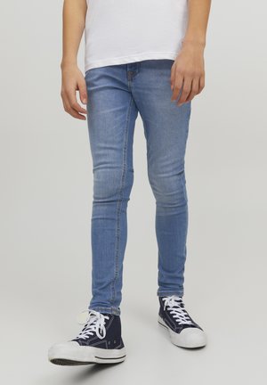 Person iført lyseblå skinny jeans og mørkeblå høje sneakers med hvide snørebånd, stående mod en ensfarvet baggrund.