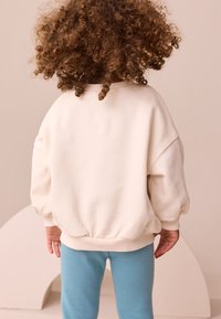 Krémová oversize mikina s žebrovaným lemem, doplněná světle modrými legínami. Textura vypadá měkce a rukávy jsou mírně nadýchané.