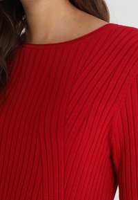 Donna che indossa un maglione a coste rosso con scollo rotondo e un sottile motivo diagonale sulla spalla.