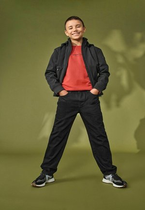 Veste zippée noire sur un t-shirt rouge, accompagnée de pantalons noirs et de baskets noires/grises ; se tient devant un fond vert.