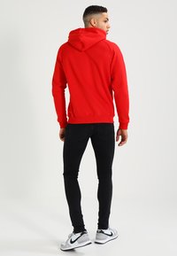 Rød hoodie med snøre i hætten, der har lange ærmer og en afslappet pasform, parret med sorte jeans og grå sneakers.
