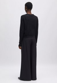 Schwarzer Strickpullover mit langen Ärmeln, geripptem Saum und weiten schwarzen Hosen aus glattem Stoff. Modell von hinten vor einem neutralen Hintergrund betrachtet.