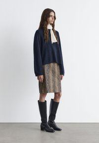 Marineblauer übergroßer Pullover mit einem cremefarbenen Halstuch, kombiniert mit einem knielangen Rock mit Schlangenprint und schwarzen kniehohen Lederstiefeln.