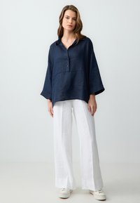 Chemise en lin bleu marine avec un col, trois boutons et des manches larges, associée à un pantalon large en lin blanc et des baskets crème.