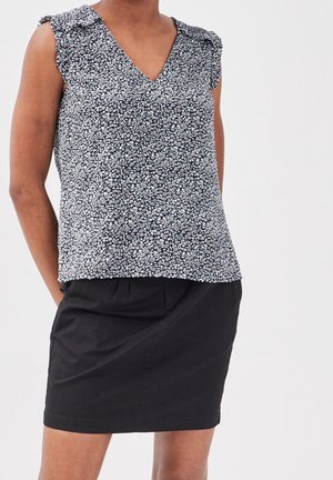 Femme portant un chemisier sans manches à motifs floraux noirs et blancs et une jupe noire unie arrivant au genou, debout les mains derrière le dos.