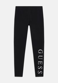 Schwarze Leggings aus elastischem Stoff, mit einem silbernen "GUESS"-Logo, das vertikal am linken Bein aufgedruckt ist. Mit elastischem Bund.