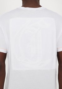 Tricou din bumbac alb cu un logo texturat reliefat pe spate, având cusături contrastante în portocaliu pe mâneci.