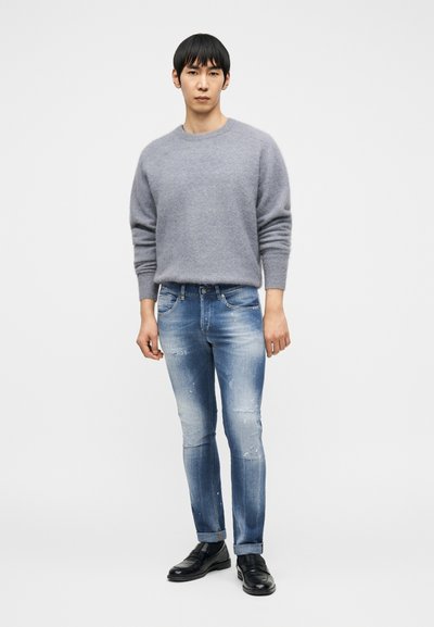 Άνδρας στέκεται φορώντας γκρι μάλλινο πουλόβερ, στενά τζιν μπλε με φθορές και μαύρα δερμάτινα παπούτσια μπροστά σε απλό φόντο.
