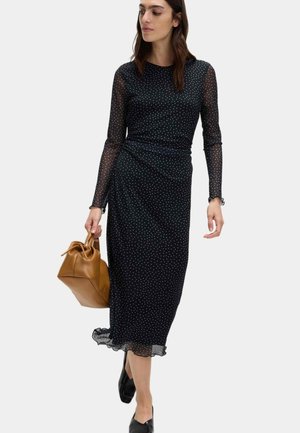 Mujer con un vestido midi de manga larga negro con lunares, sosteniendo un bolso de cuero color marrón claro y caminando hacia adelante.