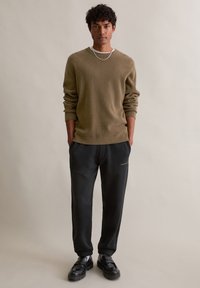 Pull à texture olive avec manches longues, associé à un pantalon noir fuselé. Des chaussures noires à semelle épaisse et une chaîne en argent complètent le look.