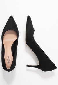 Escarpins à talons hauts en daim noir avec un bout pointu, une texture lisse et un talon fin. La semelle intérieure présente un logo embossé.