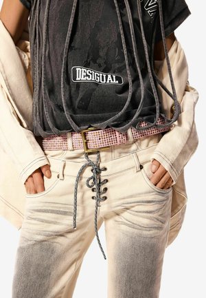 Ruke u džepovima izblijedjelih bež hlača s crnom čipkom, ružičasti remen sa zakovicama, crna majica s natpisom "Desigual" i prebačena bež jakna.