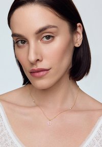 Złoty łańcuszek z małym różowym kwiatowym wisiorem i subtelnymi akcentami z koralików, noszony na nagim ramieniu, na tle w neutralnych kolorach.