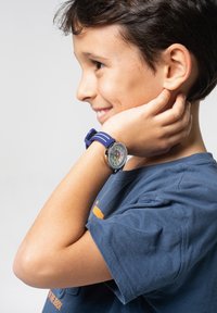 Montre analogique avec un cadran rond, des repères horaires colorés et un bracelet en silicone bleu avec des rayures blanches. Portée au poignet d'un enfant.