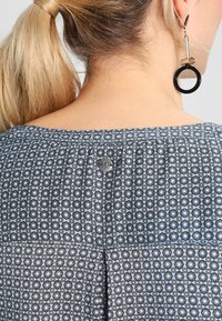 Haut bleu marine à motifs, en tissu texturé avec des motifs géométriques, un dos plissé et un détail de bouton argenté près de l'encolure.