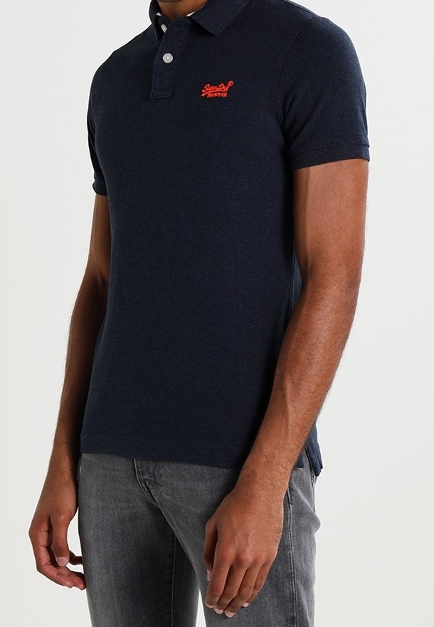 Námořnická modrá polo tričko vyrobené z bavlny, s klasickým límcem, krátkými rukávy a malou červenou nášivkou na hrudi. Hladká textura.