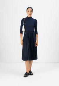 Navy pleated dress with a high neckline and three-quarter sleeves, featuring a drawstring waist. Worn with a white handbag and black loafers.