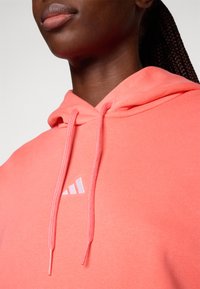 Korallfärgad hoodie med pullover-design, dragsko i huvan och vit logotypdetalj. Mjuk textur med avslappnad passform och ribbad mudd.