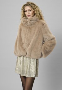 Plašč iz umetnega krzna v svetlo bež barvi z visokim ovratnikom in širokimi rokavi, v kombinaciji z bleščečo zlatimi sequini krilom. Mehka, teksturirana zunanjost.