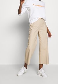 Person som bär beige vida cropped byxor, vita sneakers och en vit sweatshirt med orange text "Tommy Hilfiger".