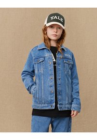 Veste en denim bleue avec poches avant, fermeture à boutons et couture contrastante, associée à un T-shirt noir et une casquette blanche et noire.