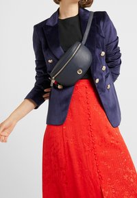 Marsupio in pelle sintetica blu con zip dorata e logo, indossato sopra un top nero e una gonna rossa brillante con texture e dettagli con bottoni.