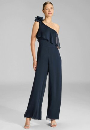 Tuta blu navy con una spalla scoperta, dettaglio asimmetrico a volant e gambe larghe. Realizzata in tessuto leggero con una texture liscia.