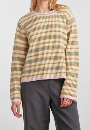 Femme portant un pull en tricot rayé pastel rose, vert et beige à manches longues et un pantalon gris foncé, debout avec les mains détendues.