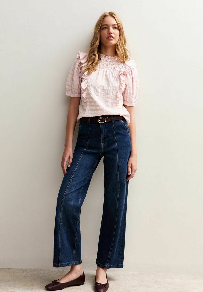 Hellrosa strukturierte Bluse mit Rüschendetails, kombiniert mit dunkelblauen weiten Jeans. Ein brauner Gürtel und braune flache Schuhe vervollständigen das Outfit.