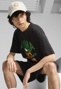 Czarna bawełniana koszulka z zielonym nadrukiem i graficznym rysunkiem; w komplecie z czarnymi szortami. Model nosi kremową czapkę z szczegółem logo.