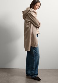 Cappotto impermeabile beige con rivestimento imbottito interno, cappuccio e vita regolabile; indossato con pantaloni in denim blu larghi e stivaletti neri.